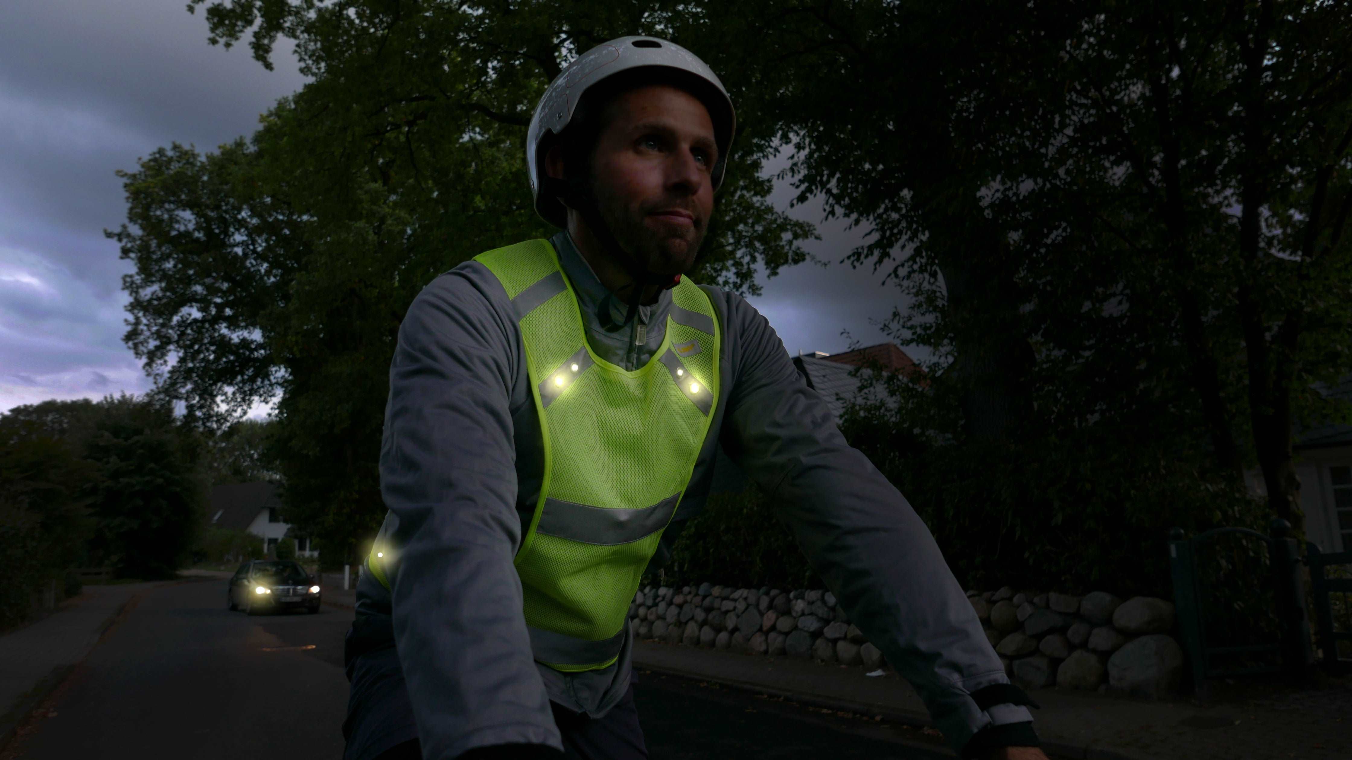 LED Sportweste  mit leuchtenden LEDs an Fahrradfahrer im Dunkeln – Bäume und Auto im Hintergrund