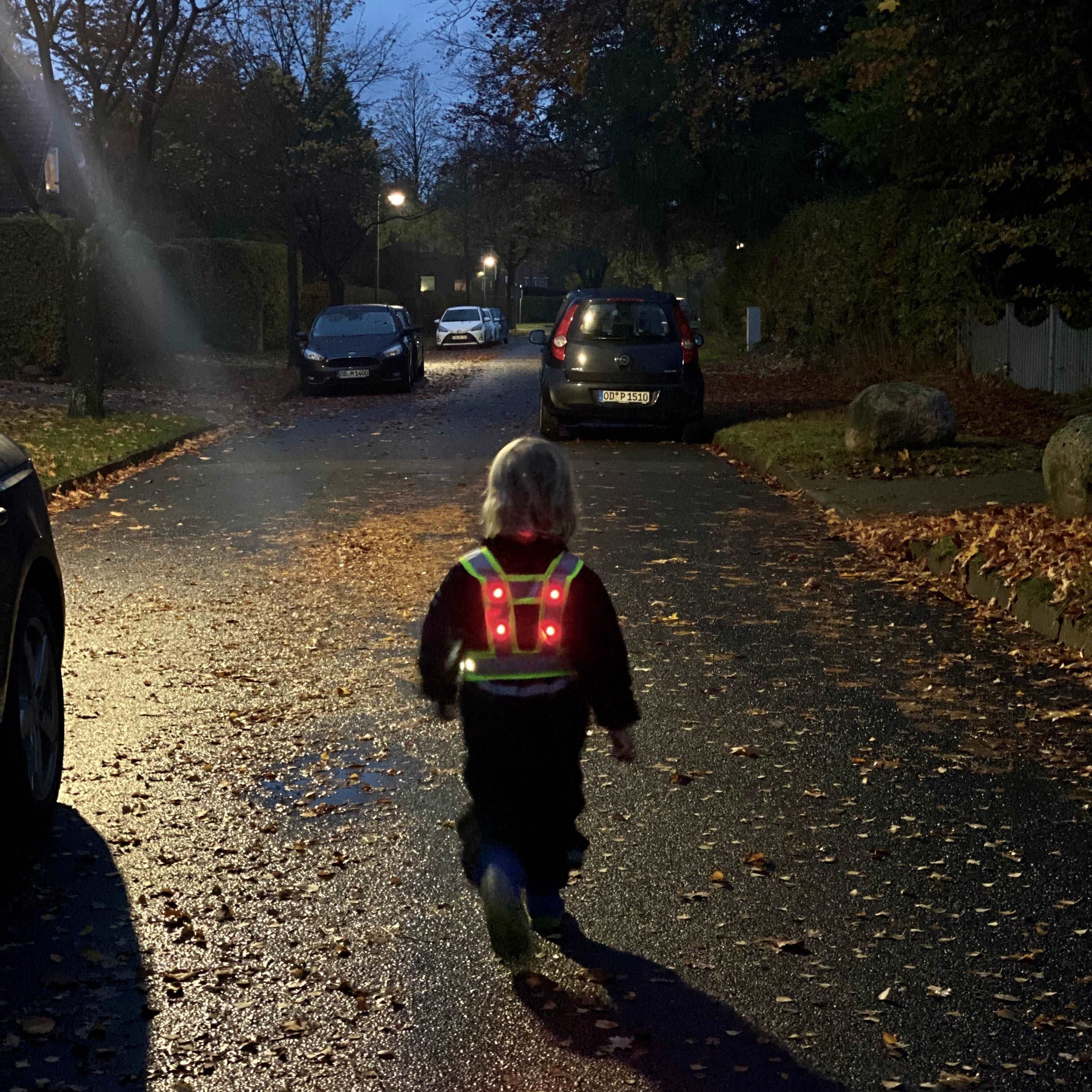 LED Warnweste Kinder mit leuchtenden LEDs an Kind im Dunkeln – Kind läuft auf Straße, parkende Autos im Hintergrund